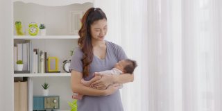 美丽的亚洲妇女抱着新生的婴儿在她的手臂站在舒适的家窗前。快乐的婴儿睡在母亲的怀里，安全舒适。母婴概念