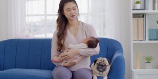 美丽的亚洲妇女抱着新生的婴儿在她的手臂坐在沙发上与可爱的哈巴狗品种在舒适的家。快乐的婴儿睡在母亲的怀里，安全舒适。母婴概念