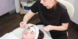 美容师在美容诊所、水疗沙龙用化妆刷给年轻漂亮的女性脸上涂面膜