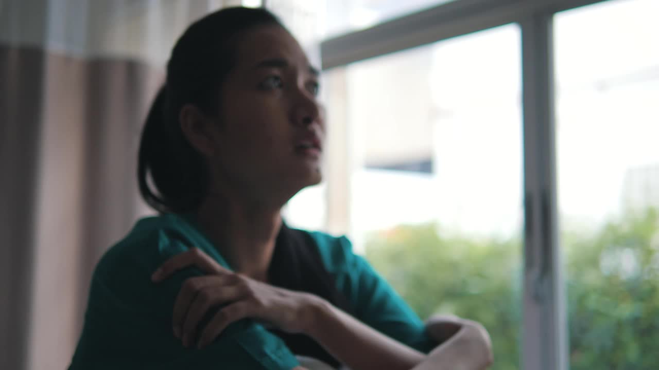 亚洲妇女坐在她的床上和哭泣在医院的医疗长袍。她是一个因头痛住在医院病房里的沮丧沮丧的病人。