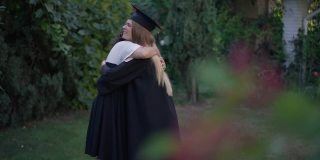 快乐年轻的女毕业生拥抱着成熟的母亲在户外的花园。毕业典礼那天，微笑苗条美丽的女儿在后院拥抱父母。教育与家庭观念