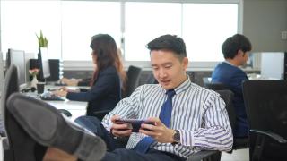 懒惰的亚洲商人玩着游戏打发工作时间在办公室打电话，同事履行职责视频素材模板下载