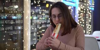 一位戴眼镜的年轻女子在餐馆里吃着墨西哥卷饼。晚上，一个女孩坐在装饰着花环的咖啡厅里。