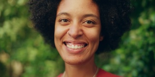 一个快乐的年轻拉丁人的近距离肖像与棕色的眼睛，非洲式的头发和鼻子穿孔为相机摆姿势。美丽多样的多民族黑人西班牙裔女性微笑在绿色的自然背景。