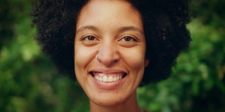 一个快乐的年轻拉丁人的近距离肖像与棕色的眼睛，非洲式的头发和鼻子穿孔为相机摆姿势。美丽多样的多民族黑人西班牙裔女性微笑在绿色的自然背景。