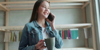 亚洲女性创意设计师项目通过智能手机对话与客户进行讨论和展示，亚洲休闲服饰放松与客户或朋友交谈，手握智能手机和热咖啡，站在家庭工作室的工作台前