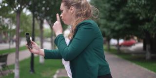 侧视图肖像愤怒的女博主手势说话的智能手机站在城市的街道上。不满的白人胖女人抱怨博客在线直播。