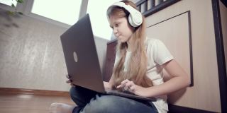 一个聪明的七岁小女孩戴着白色耳机，手里拿着笔记本电脑，在她房间的地板上推着。年轻一代对互联网和IT技术很感兴趣