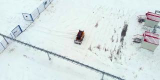 一辆小型拖拉机装载机清理石油和天然气设备建筑附近的积雪。一架无人机在工业区内被雪覆盖的石油工厂和燃气轮机发电厂上空飞过