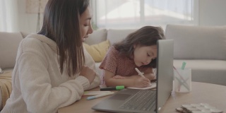 亚洲母亲帮助学龄前的女儿女孩在家里使用笔记本电脑电子学习，放大在线虚拟课堂，家庭教育，新常态的概念