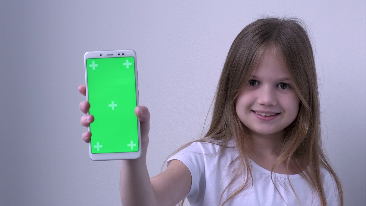 可爱的小女孩用绿色的屏幕展示手机，模拟手机，手机。绿屏智能手机的色度键设置为广告。科技、小玩意和孩子。儿童应用演示