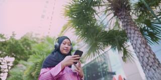 亚洲迷人的穆斯林女子戴着耳机一边听音乐一边拿着咖啡杯走在现代市中心的街道路边，一天，蓝牙连接设备，生活娱乐