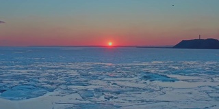 从空中俯瞰冬天冰冻海面上的日落。无人机向前飞向日出和地平线。直升机飞越融化的冰层，向太阳漂移。春天变暖。生动的色彩斑斓的天空