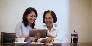 亚洲女儿和老年母亲享受在咖啡休息和看数字平板电脑，而坐在家里的桌子上