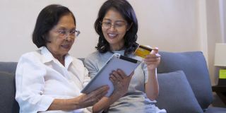 亚洲女儿和老年母亲在家里客厅的沙发上使用信用卡和数字平板电脑支付