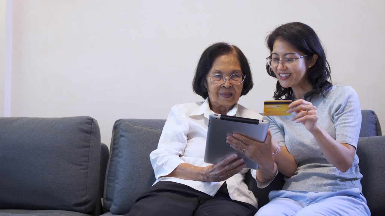亚洲女儿和老年母亲在家里客厅的沙发上使用信用卡和数字平板电脑支付