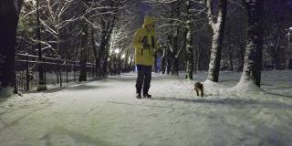 被训练用皮带走路的猫。傍晚，一个穿着黄色夹克的女人带着她的猫走在一个冬季公园的雪道上。