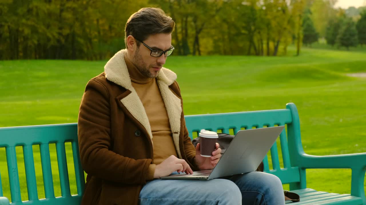 戴眼镜的商人坐在公园里，喝着咖啡，拿着笔记本电脑