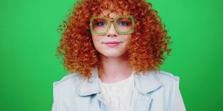 年轻俏皮的红发卷发女人向相机吹五彩纸屑，微笑着越过绿色的色度键背景