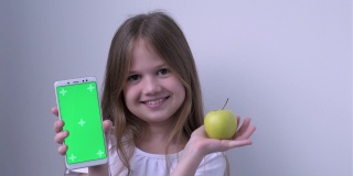 可爱的小女孩用绿色屏幕展示苹果和手机模拟手机、手机、手机。绿屏智能手机的色度键用于广告。水果、健康饮食、儿童口腔保健
