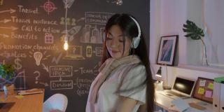 无忧无虑的亚洲女商人戴着耳机在晚上的创意办公室跳舞