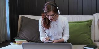 大笑的年轻女子戴着耳机会议，在笔记本电脑上打电话与在线教师学习，工作在家里。