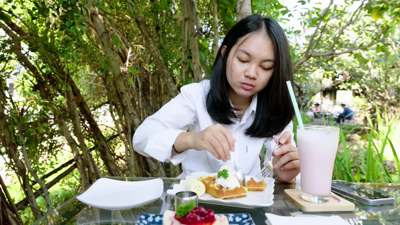 4k，公园里，一名身穿白衬衫的年轻亚洲女子正在香蕉华夫饼上舀冰淇淋。桌上还有草莓奶昔