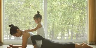 4 k。亚洲妈妈在家里练习瑜伽，有一个可爱的女儿坐在妈妈背上。隔离自我隔离期间的培训。休闲健康的生活方式。健康的家庭在早晨的活动