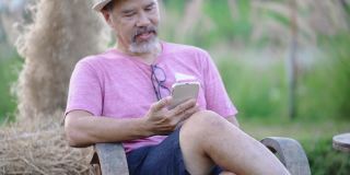 笑容满面的亚洲灰胡子成熟男子坐在户外的花圃里，用手机上的在线约会app聊天。快乐的退休男人穿着粉色衬衫，戴着眼镜，日落时戴着帽子。普通情人节新概念