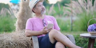 笑容满面的亚洲灰胡子成熟男子坐在户外的花圃里，用手机上的在线约会app聊天。快乐的退休男人穿着粉色衬衫，戴着眼镜，日落时戴着帽子。普通情人节新概念