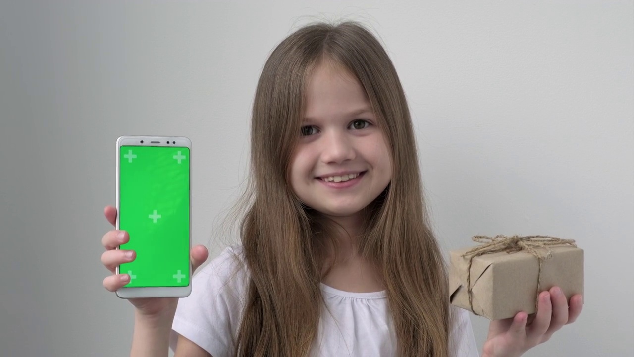 可爱的小女孩用绿色屏幕展示手机模拟手机、手机、电话和礼物。绿屏智能手机的色度键设置为广告。圣诞节，情人节，爱，母亲节