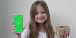 可爱的小女孩用绿色屏幕展示手机模拟手机、手机、电话和礼物。绿屏智能手机的色度键设置为广告。圣诞节，情人节，爱，母亲节