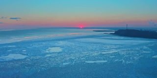 从空中俯瞰冰冻的冬季海面上的日落。一架无人机在日出时降落在海面上。在融化的浮冰和工业城市上空飞行。全球变暖。生动的色彩斑斓的天空。工业厂房