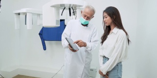 戴着防护面罩的老牙医在x光室里向一位亚洲中年女病人解释用药片拍的x光片，她仔细地听着