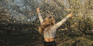 穿着毛衣、牛仔裤的迷人年轻女子在盛开的苹果树间翩翩起舞。日落，春天的果园和绿色的田野。慢动作