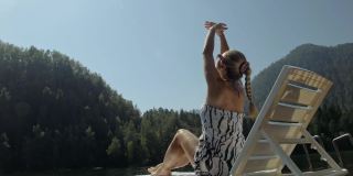一个女人戴着太阳镜，披着波西米亚风格的丝绸披肩，躺在日光浴床上。女孩在水下的泛木码头休息。人行道上覆盖着湖水