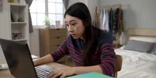 这是在现代风格的卧室里，坐在书桌前的电脑上学习的亚裔女大学生开心地看着镜头的照片。