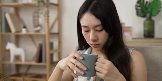 独自呆在家里的亚洲女性在喝茶，脸上露出忧虑的表情。