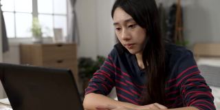 焦急的亚洲女学生用手指敲着桌子，在家里用电脑学习的过程中，想到了解决问题的好办法，露出了笑容
