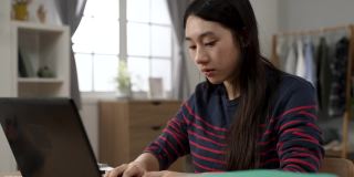 一名快乐的亚裔女学生一边在书桌前的笔记本电脑上输入成绩单，一边对着镜头微笑着转着头。