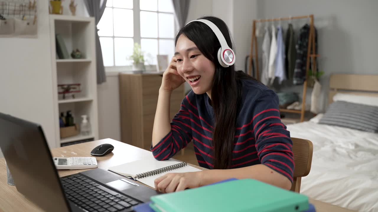 一名亚洲女学生戴着头戴式耳机，面带微笑地跟着老师重复着在家里用电脑学习语言的视频。