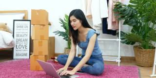 4 k。一名亚洲妇女在卧室地板上的笔记本电脑上打字，手里拿着包裹盒。电子商务，网上购物，网上零售时装店，小企业主概念