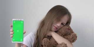 可爱的小女孩展示着绿屏手机，拥抱着泰迪熊。绿屏智能手机的色度键设置为广告。促销玩具商店。圣诞节，情人节，爱，母亲节
