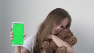 可爱的小女孩展示着绿屏手机，拥抱着泰迪熊。绿屏智能手机的色度键设置为广告。促销玩具商店。圣诞节，情人节，爱，母亲节视频素材模板下载
