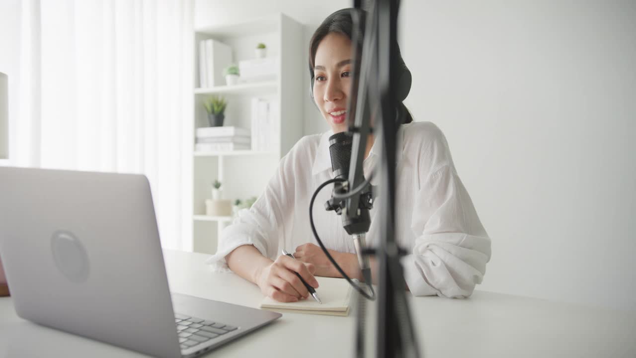 年轻的亚洲妇女在她的笔记本电脑从家庭工作室录制播客。