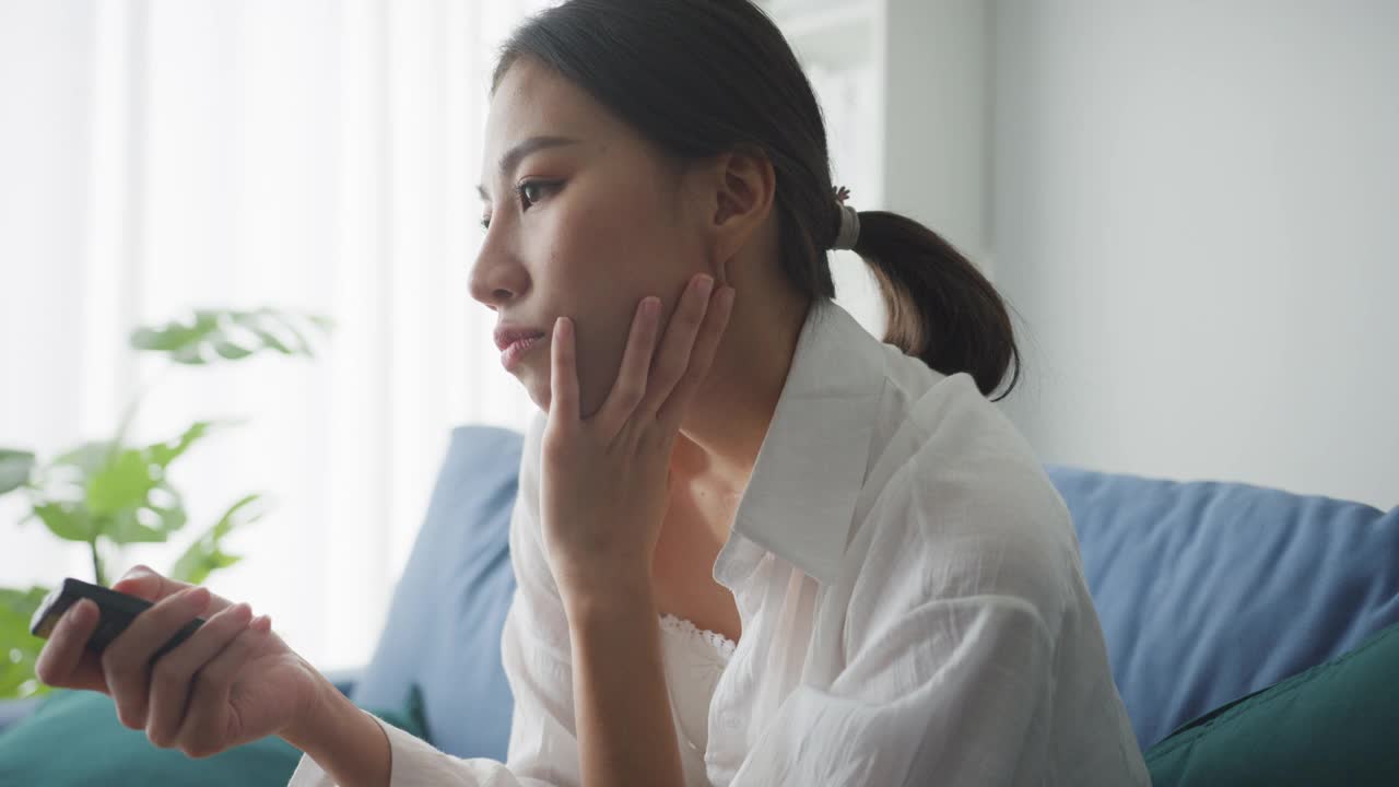 无聊的年轻亚洲女人坐在客厅沙发上看电视在家里。