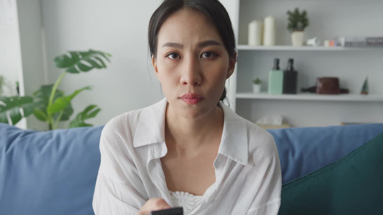 年轻的亚洲妇女坐在家里客厅的沙发上看电视。