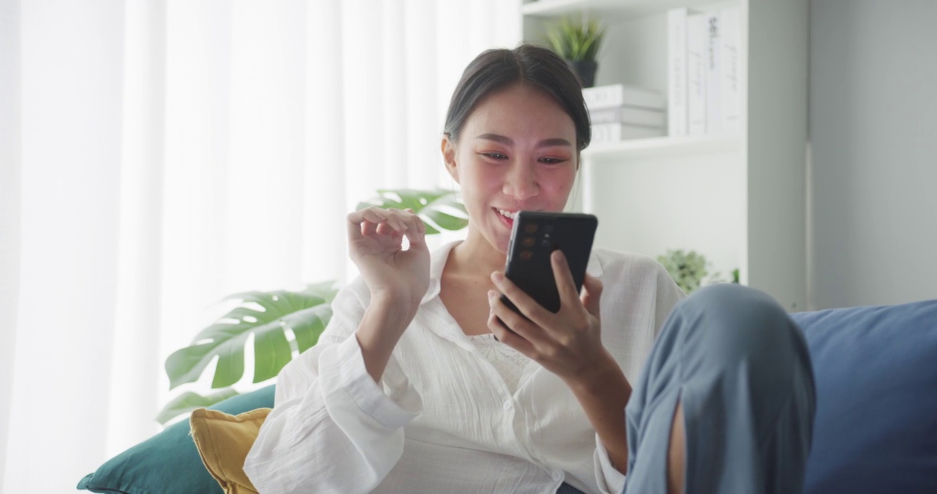 年轻的亚洲女性坐在家里客厅的沙发上使用智能手机。