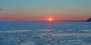 从空中俯瞰冰冻的冬季海面上的日落。无人机向后飞向日出和地平线。直升飞机在融化的冰层上飞向太阳。春天变暖。生动的色彩斑斓的天空