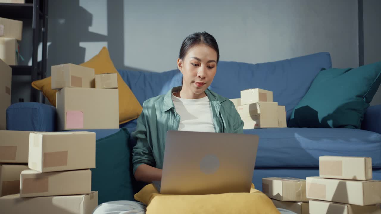 年轻的亚洲女售货员在网上商店使用笔记本电脑准备包裹发货给客户在家里。